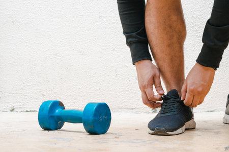 man tying his shoe standing next to blue dumbbell, before exercising. exercise at home, white...
