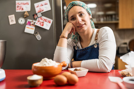 Portrait of a latin adult woman cook sitting in the kitchen with her right hand in her cheek smiling and looking away with some eggs, flour, and other ingredients for the preparation of homemade argentine alfajores.の写真素材