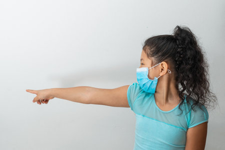 little girl (8 years old) sitting with her right hand pointing to the right, brown girl teasing another person with a blue surgical mask. Medical, pharmaceutical and sanitary concept.の写真素材