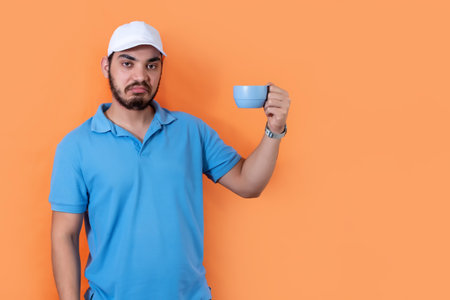 Dissatisfied man. Bearded guy holds mug of hot drink, on orange background.の写真素材