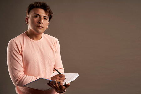 latin man writing in notebook standing facing front isolated on gray background with pink clothes copy spaceの写真素材