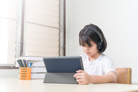 Cheerful Asian little girl student wears headphone, writes on digital tablet to study at home. Education Concept Stock Photoの写真素材