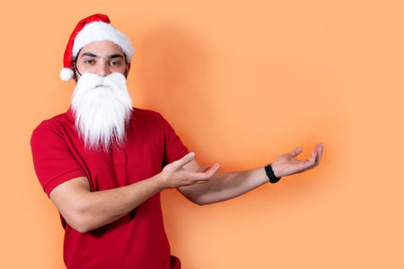 Young man with Santa Claus hat and white beard, pointing with hands at wall. Front view.の写真素材