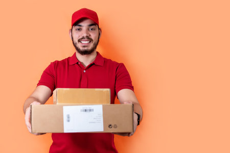 Delivery man, in red uniform, holding several orders, looking at camera, on orange background.の写真素材