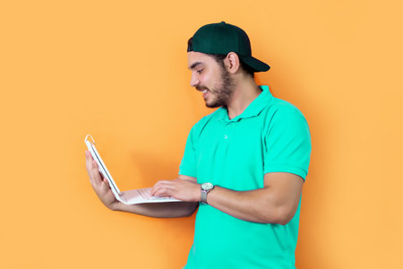 Young man with beard, T-shirt and green cap, using a tablet with keyboard, on yellow background.の写真素材