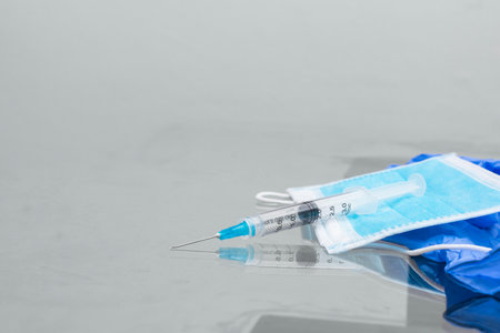 syringe on top of blue protective mask and blue rubber gloves, on top of glass table, ready to be used for vaccination. instruments for vaccination (covid-19, influenza) pandemic and health conceptの写真素材