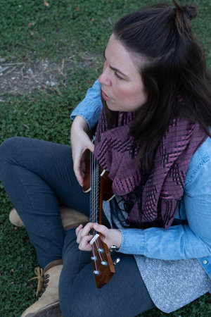 young brunette girl with long hair playing ukulele in the field on the grass