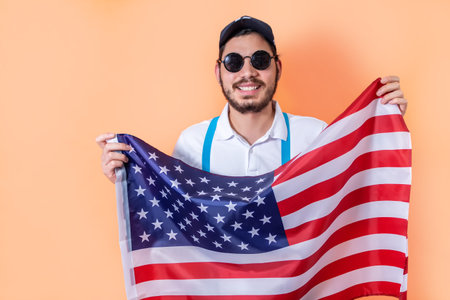 Happy young hipster holding USA flag, on light orange background.