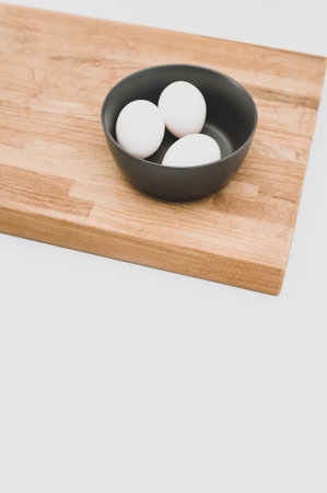organic white eggs in a dark gray bowl and wooden chopping board on a white surface with copy space...
