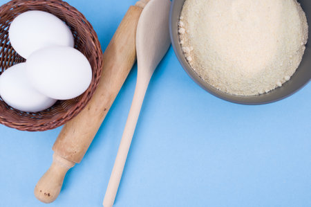 wooden rolling pin white eggs and dark gray bowl with wholegrain flour on blue background with copy spaceの写真素材