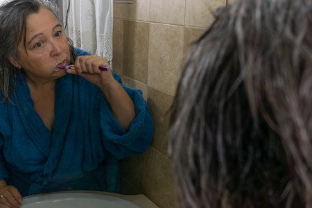 mature woman brushing her teeth in front of a mirror in her bathroom at homeの写真素材