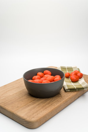 dark gray bowl full of datterini tomatoes with chopping board and table napkin on a white surface with text spaceの写真素材