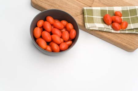 dark gray bowl full of datterini tomatoes with chopping board and table napkin on a white surface with text spaceの写真素材