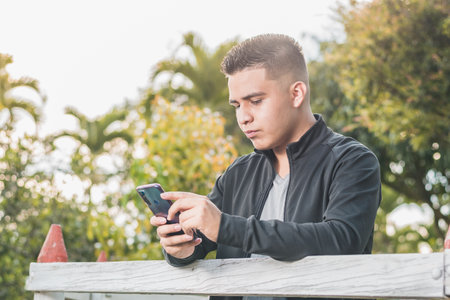 Young businessman looking at his social networks on his cell phone with a serious look and very focused on the business and his accounts, next to a white picket fence in an outdoor parkの写真素材