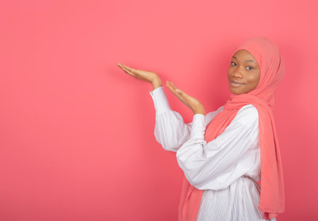 Young religious Muslim woman raises her palms over the blank, holds an invisible object, wears a pink silk scarf on her head, promotes the article.の写真素材