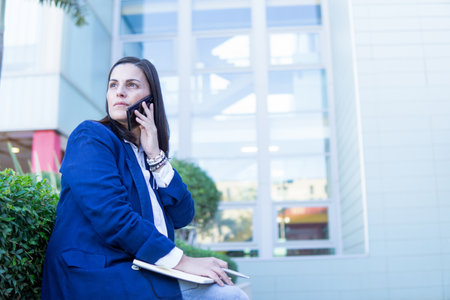 Beautiful Caucasian businesswoman calling phone and holding a notebook outside an office building. She is wearing a casual blue jacket.の写真素材