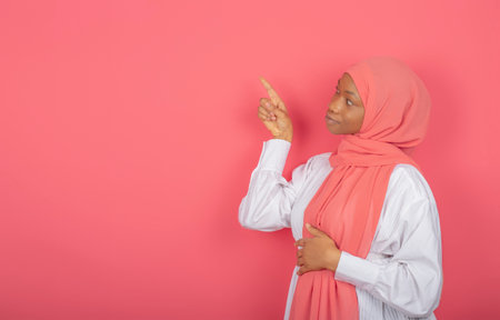Photo of young healthy muslim woman points index finger to free space, has no makeup, wrapped in pink veil isolated on pink background.の写真素材