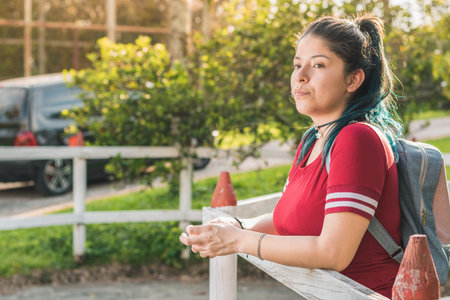 Beautiful young Latina college girl, waiting for a person next to a wooden fence, dressed in red and with blue dyed hair, with a beautiful sunset in the background and a car driving away.の写真素材