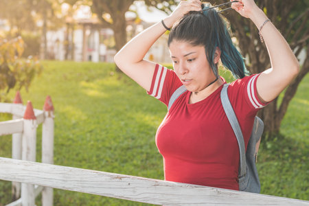 Beautiful young Latina college girl, tying her hair in a ponytail, dressed in red and dyed blue hair, standing next to a white fence, with a beautiful sunset in the background. concept of freedom.の写真素材