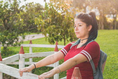 Beautiful young woman student, holding back with her two hands on a wooden fence, very happy for her life project, dressed in red with blue dyed hair, with a beautiful sunset in the background.の写真素材