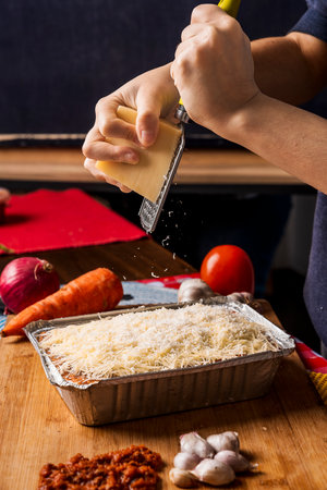Vertical image of an unrecognizable person grating fresh cheese over lasagna with a grater with different types of ingredients around the food.の写真素材