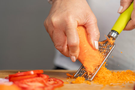 Close up view of the hands of an adult woman grating a carrot with a grater on a wooden board for lasagna preparation with copy space.の写真素材