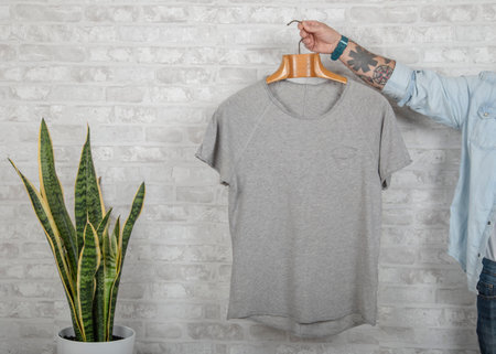 Tattooed male hand holds a hanger with gray top quality fine cotton t-shirt. Mockup Copy Paste Adの写真素材