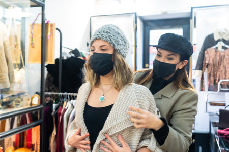 Employee with a mask in a clothing store trying on a jewel to a client, working with security...