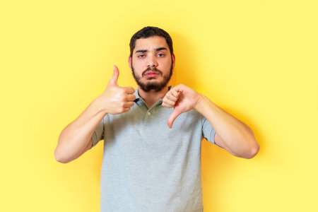 Confused bearded man is showing both like and dislike gestures over yellow background. Undecided between yes or not.の写真素材