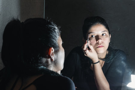 beautiful latina woman in front of the mirror making up her eyes. girl looking at herself in the mirror painting her eyebrows at home with a makeup brush. beauty, cosmetics and handicraft concept.の写真素材