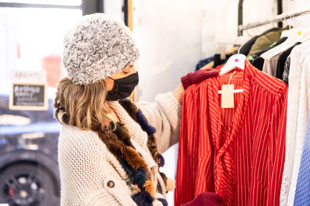 A customer with a protective mask looking at new collection clothes on a hanger, second-hand and sustainable clothing store, coronavirus, covid-19の写真素材