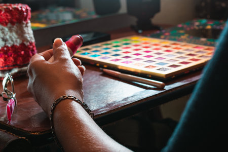 view of a girl's arm holding a lipstick in her hand, next to her brown wooden dressing table, with her make-up on top. palette of eye shadows with make-up brush and brush on a brown table.の写真素材