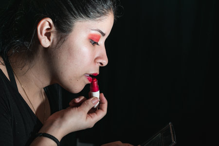latina woman using a lipstick to paint her lips red, opening her mouth a little to apply the lipstick. her eyes are painted with a red shadow. black background. looking at herself in the mirror.の写真素材