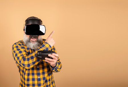 gamer in VR glasses on a yellow background holds a device and points his fingers to a place for text, a man with wireless gadgets for virtual realityの写真素材