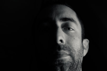 Dramatic portrait of a young man half expressionless face in deep shadow looking at camera isolated on black background.の写真素材