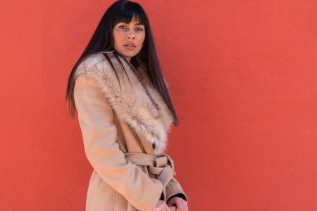 Posing of a smiling brunette model in a winter jacket leaning against a wall with a bag. Caucasian pretty a red backgroundの写真素材