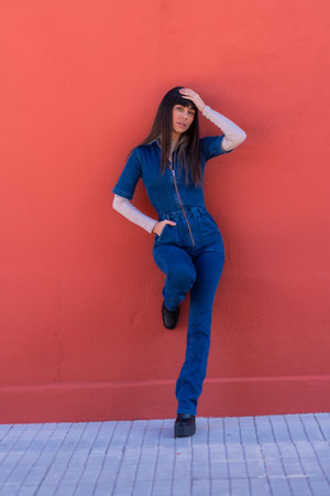 Posing of a smiling brunette girl leaning against a wall in a blue denim outfit. Pretty Caucasian posing on a red backgroundの写真素材