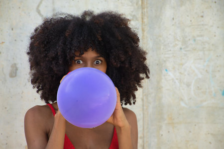 Young, beautiful African American woman inflating a purple balloon on a gray cement background.