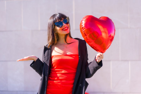 Brunette caucasian girl with a heart balloon on Valentine's day, sunglasses and red dress, Lifestyle...