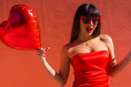 Caucasian brunette girl with heart balloons on Valentine's day wearing sunglasses, red background. Enjoying the day of celebrationの写真素材
