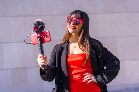 Brunette caucasian girl making a vlog on Valentine's day for social networks, sunglasses with red hearts, phone and microphone on a gray backgroundの写真素材