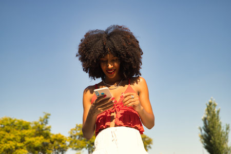 Young beautiful afro american woman with afro hair and coral top checking social networks on her cell phone.の写真素材