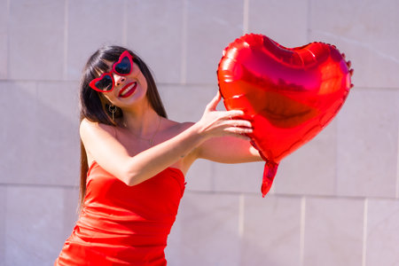 Brunette caucasian girl in love on Valentine's day with a heart balloon, sunglasses and red dressの写真素材