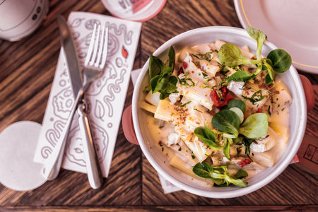 pasta with sauce, cheese and basil in a white dish on a wooden table in a restaurant, a rowl knife...