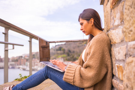 A young Latin woman in a white t-shirt and a brown wool sweater working on the terrace of her house by the seaの写真素材
