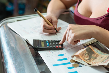 woman reviewing her business statistics chart. balancing sales while counting money. writing with an eco-friendly cardboard pen and a small black calculator on a large notebook.の写真素材