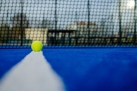 Selective focus. Ground level view of a ball on the line of a blue paddle tennis court with the net out of focus in the background. Racke sportsの写真素材