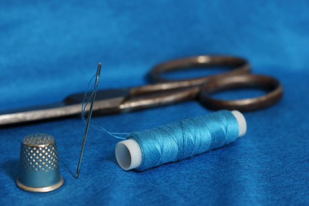 Spool of Blue Thread, Thimble and Needle on Blue Fabric With Vintage Scissors in the Background