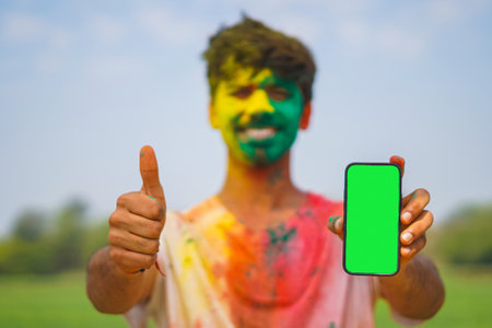 Young Indian boy covered in holi colors holding a mobile phone with green screen showing thumbs up. Holi festival and technology concept.の写真素材