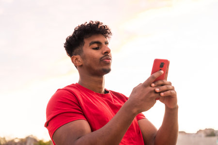 Arab young man with red t-shirt in a sunset typing a message in the cityの写真素材